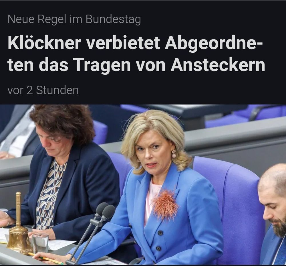 Das Bild zeigt eine Szene aus dem Deutschen Bundestag. Im Vordergrund sitzt Julia Klöckner (CDU), die aktuell das Amt der Bundestagsvizepräsidentin innehat. Sie trägt einen auffälligen blauen Blazer mit einer großen, federartigen Brosche (Anstecker) auf der linken Seite – ein ironisches Detail im Kontext der Schlagzeile.

Links neben ihr sitzt eine Frau, offenbar vertieft in ihre Unterlagen, rechts ein Mann, ebenfalls mit konzentriertem Blick nach unten. Auf dem Tisch vor Klöckner stehen zwei Mikrofone, ein Wasserglas, ein goldener Parlamentshammer und einige Papiere – typische Utensilien für eine Bundestagssitzung.

Die Schlagzeile darüber lautet: „Neue Regel im Bundestag: Klöckner verbietet Abgeordneten das Tragen von Ansteckern“

Ironie und Widerspruch entstehen durch den Kontrast zwischen dem Verbot und Klöckners eigenem auffälligen Anstecker, was auf humorvolle oder kritische Weise interpretiert werden kann.

