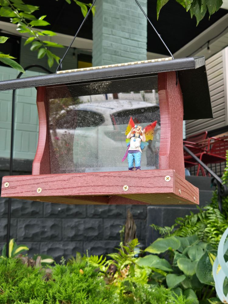 Photo of a wooden bird feeder. Along one edge someone has perched a little figurine of someone with blue hair and rainbow butterfly wings. 