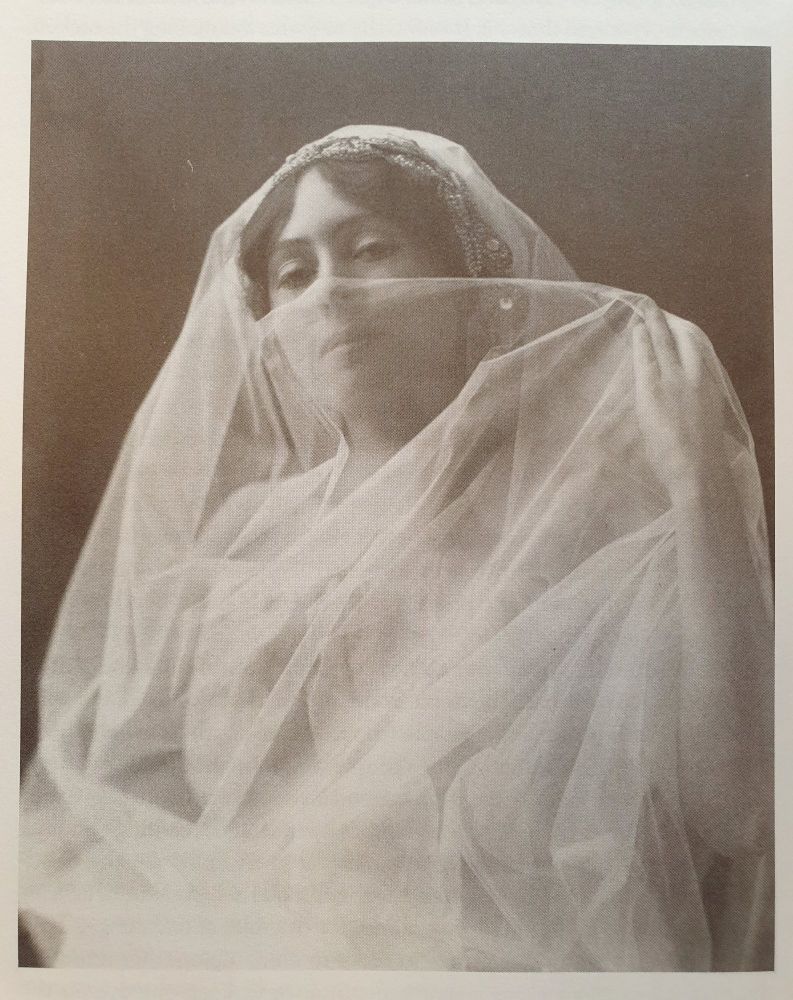 Image of Neith Boyce as a young woman staring directly at the viewer. She is shrouded in a transparent white veil which she lifts across her face.