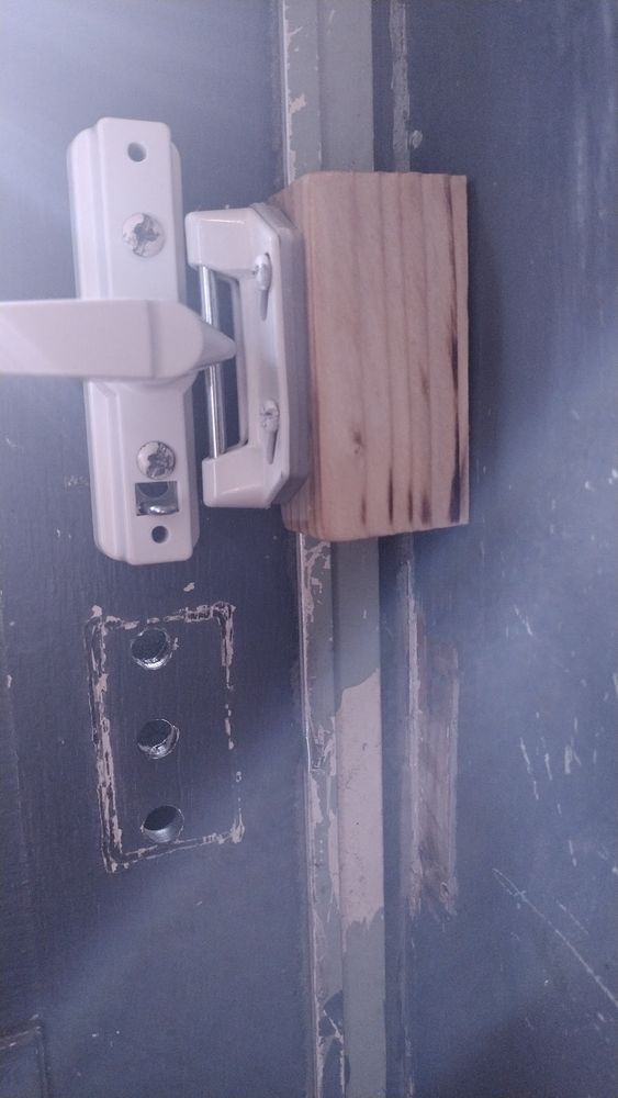 The image shows a close-up view of a door latch mechanism. There is a white metal latch attached to a piece of light-colored wood, which in turn is mounted on a doorframe. The wood appears to be cut roughly, with visible grain patterns. Two screws are securing the latch to the wood. On the left side, there is a section of a dark-colored door with chipped paint, revealing lighter colors beneath. The door has two visible holes, one above the other, where a previous latch might have been installed.
