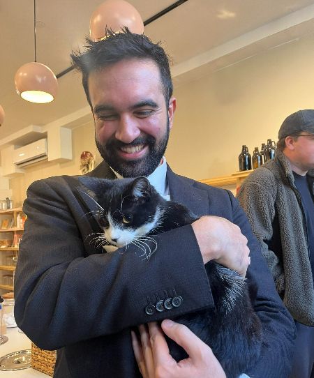 The image shows a person holding a black and white cat in an indoor setting. The person is wearing a dark jacket and smiling, with closed eyes, appearing content as they hug the cat. The cat, with a mostly black coat and a white patch on its nose and chest, is being gently held and seems relaxed. In the background, there are wooden shelves with bottles, and overhead are spherical pendant lights with a soft orange hue.