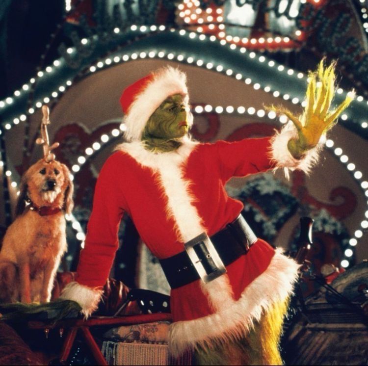 grinch in the santa suit with his arm outstretched and fingers out straight. you can see max the dog sitting behind him. as well as several christmas lights.