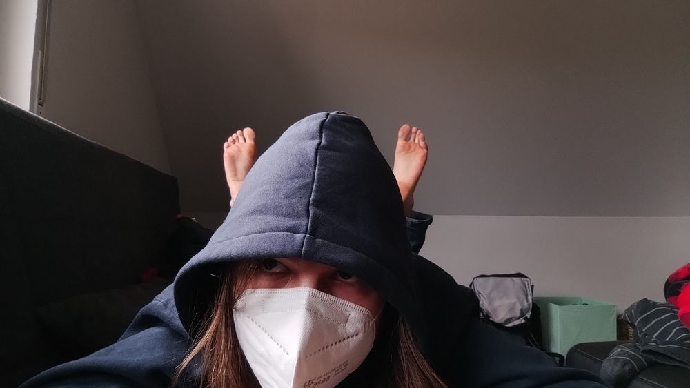 Photograph of me lying on my belly on the sofa, wearing a facemask and dark blue hoodie. I am kicking my bare feet up behind me.