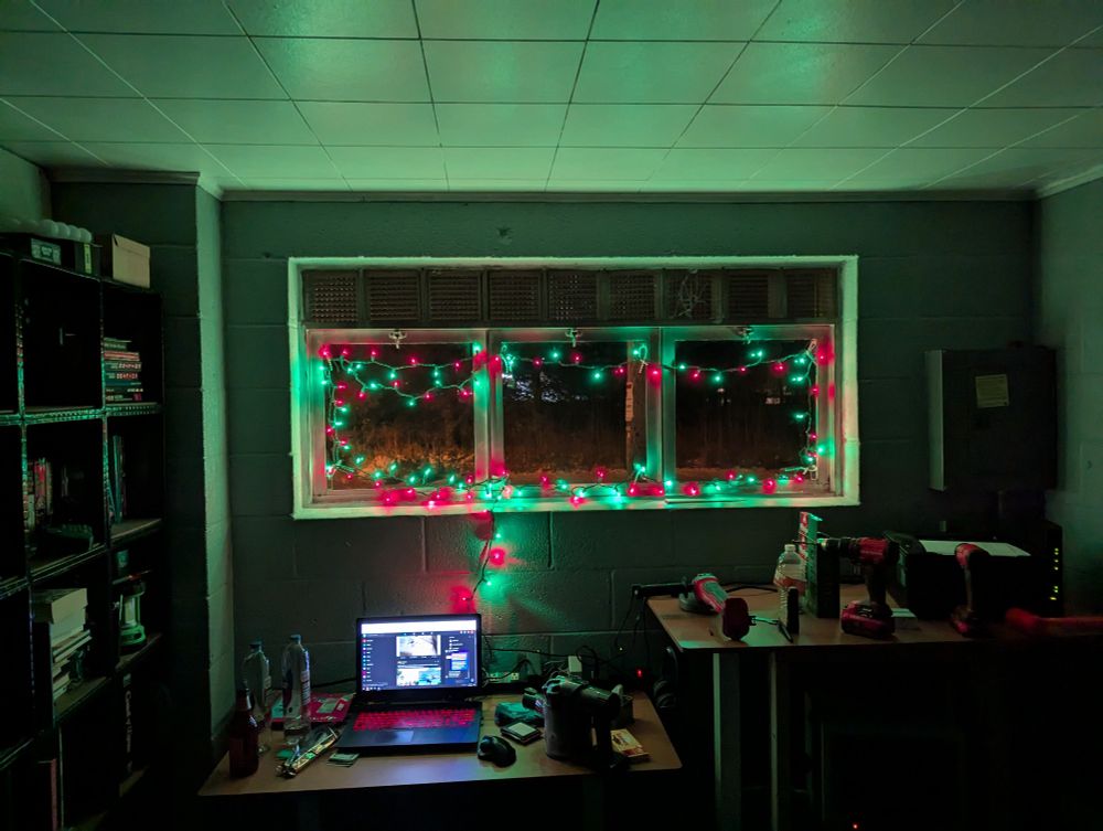 A picture of a messy office with some Xmas lights around the window to bring some holiday cheer.