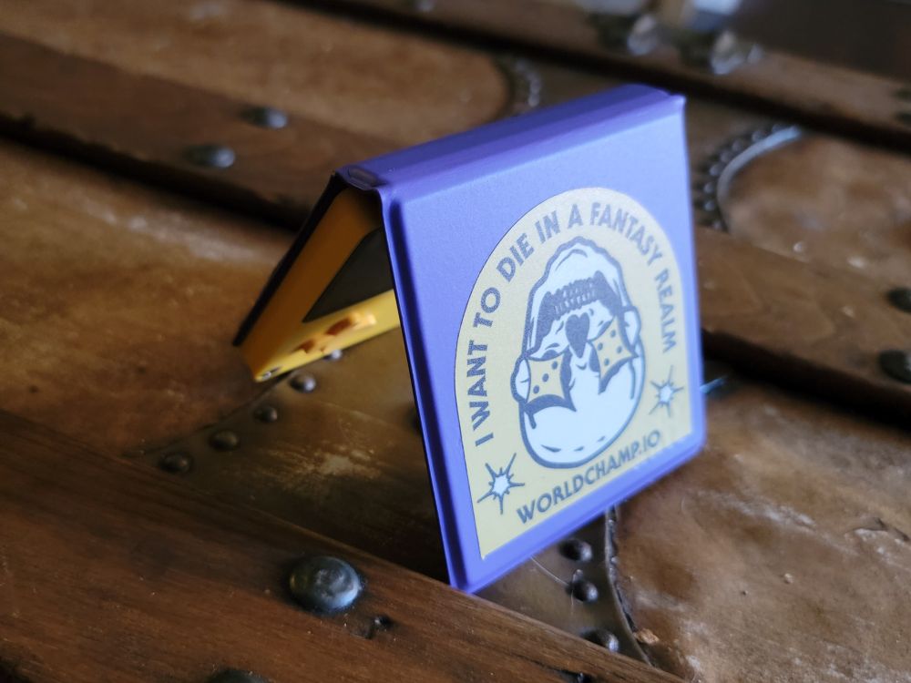 My Playdate — a small yellow handheld game device, like a Gameboy with a crank — with its purple cover proping it upright. There's a yellow World Champ Games sticker on the cover with an upside-down skull, dice (d6 showing 3 and 4) for eyes and grinning. The banner text says "I WANT TO DIE IN A FANTASY REALM".