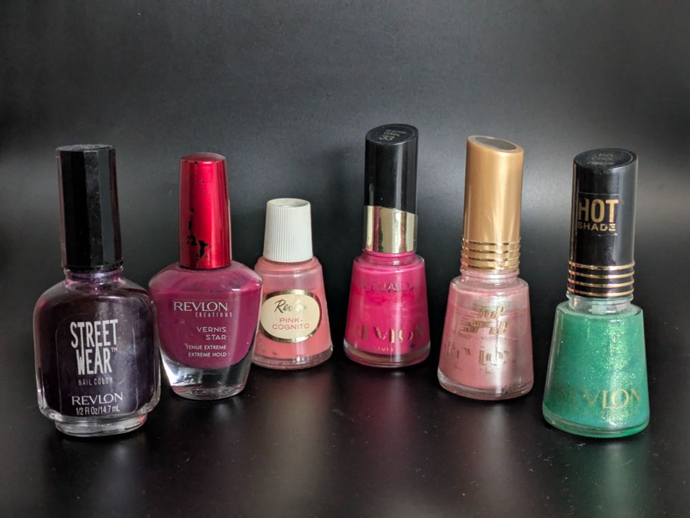 Six different types of Revlon Nail polish. A dark purple Street Wear, a mauve Vernis Star, a bottle from the 50s called Pink Cognito, a bright pink eighties bottle, a pink shiny bottle with a tan cap and a green glittery bottle with "Hot Shade" on the cap