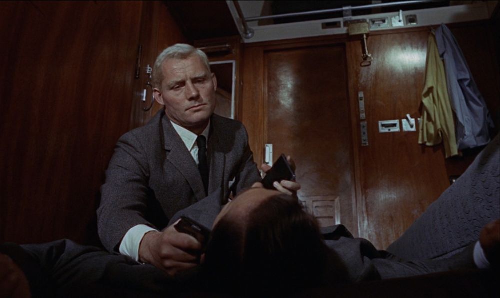 A man kneels over Bond on the floor of a railway carriage. In his right hand he holds a gun to Bond's head. In his left hand he holds a cigarette case.