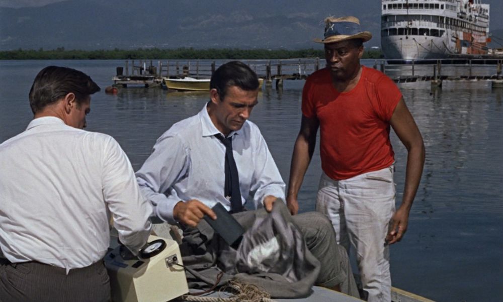 James Bond is sitting on a boat. A man stands to the right of him, while another sits to the left of him, with his back to the camera. He is taking a cigarette case out of his jacket, which he is not wearing.