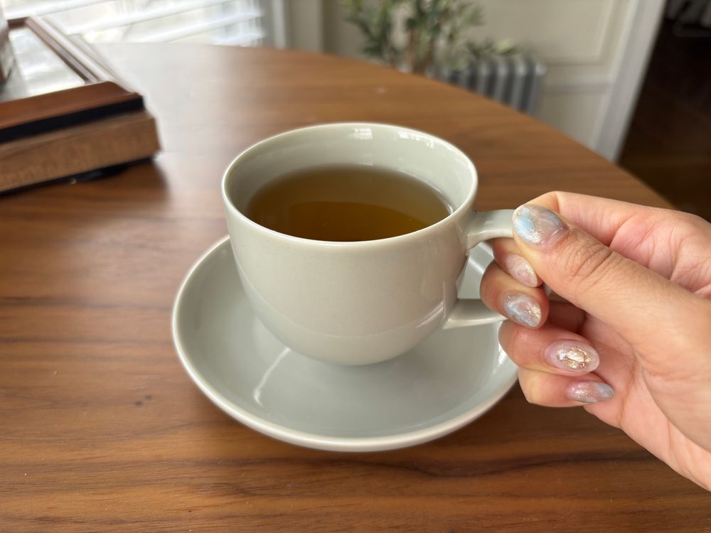 Cozy and serene photo of a hand holding a light grey thin-lipped tea cup filled with jasmine tea. Below the cup is a matching coaster. 