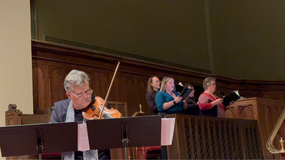 Mid-performance: Peter with bow and violin, reading music of two stands; behind him is half of the Choir of St David’s.