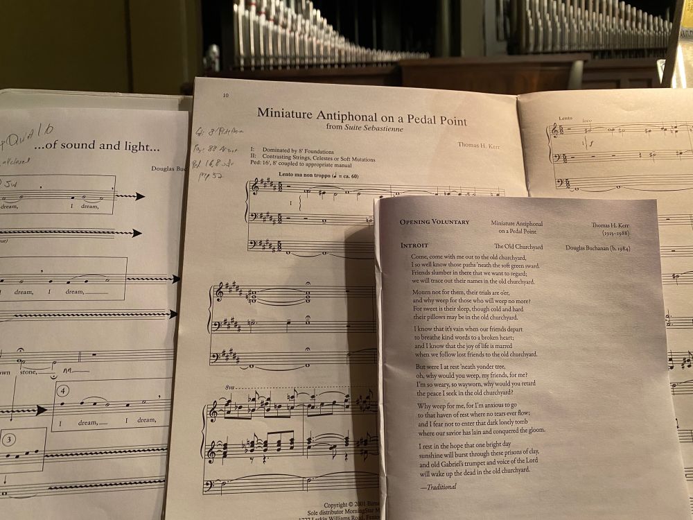 ID: Sheet music on a music rack, looking up at the pipes of a pipe organ.