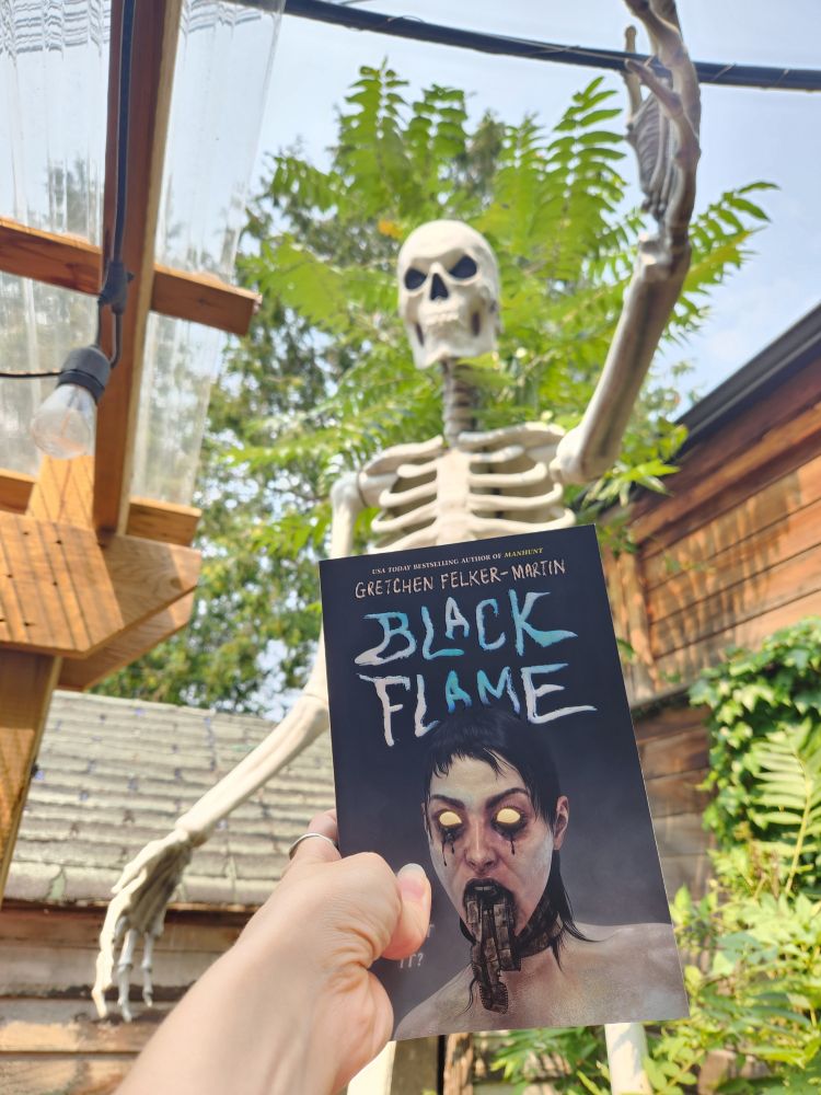 The book Black Flame by Gretchen Felker Martin held in front of a 10 foot tall plastic skeleton outside on a patio