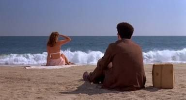 A still from Barton Fink (Dir. Joel and Ethan Coen, 1991). In the foreground, Barton sits in a dark wool suit, a cardboard parcel beside him, facing the ocean. In the foreground a blonde woman sits just in front of a breaking wave and raises her hand to her eyes.