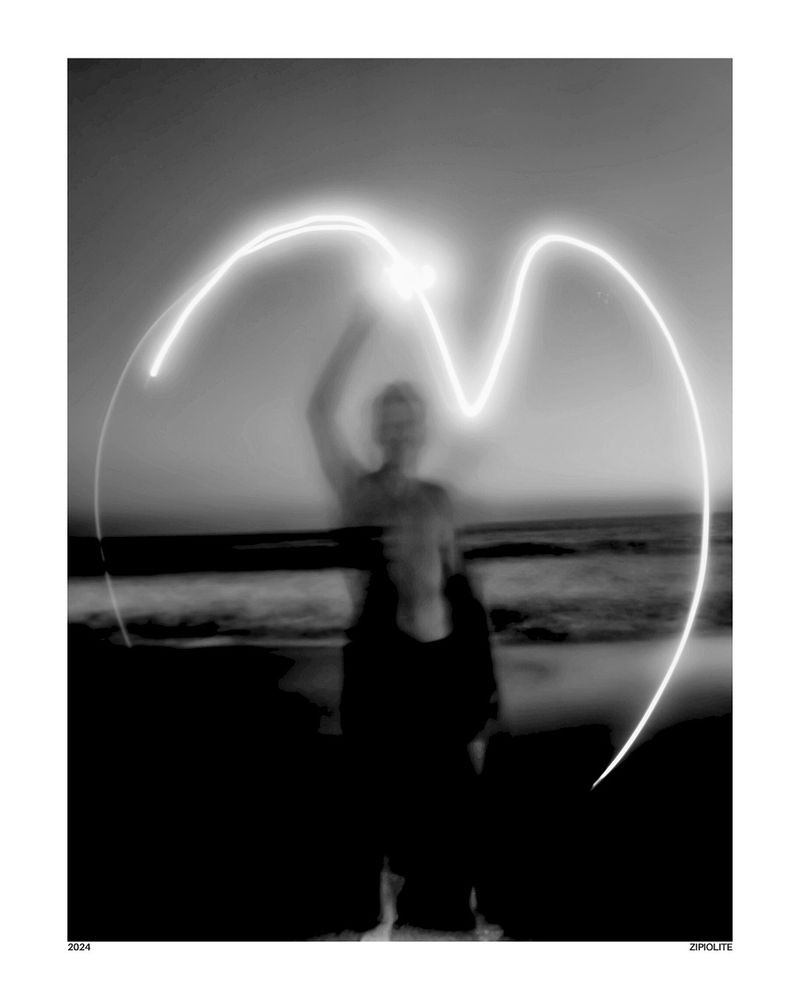 A black and white long exposure photograph of a person on a beach at dusk, using a light source to draw a luminous, heart shape in the air around them. The ocean waves are blurred in the background.