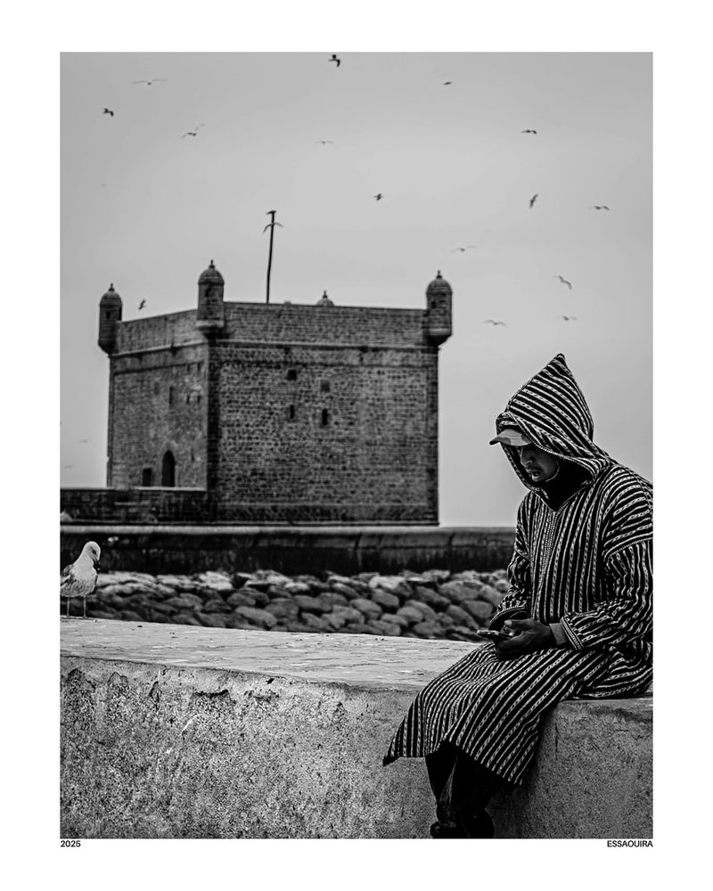 A young man in a striped djellaba sits alone on a ledge in Essaouira, Morocco. His head is down, focused on his phone, while behind him rise ancient stone towers and fortress walls. The scene is quiet, the contrast between centuries-old architecture and modern technology subtly emphasized in black and white.