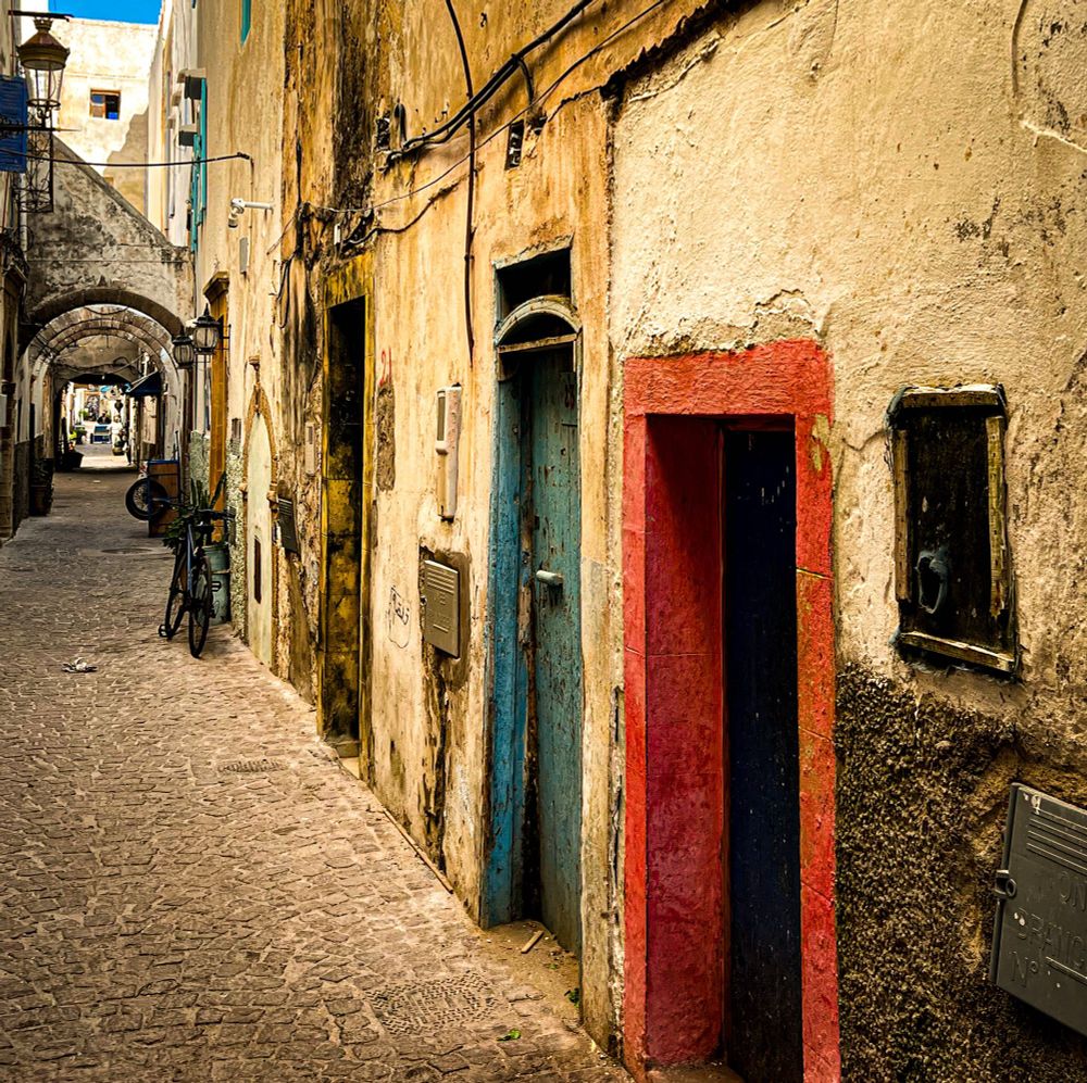 A narrow cobblestone alley in the Essaouira medina, lined with weathered, colorful doors and textured, peeling walls. A lone bicycle leans on the wall under a stone arch.
