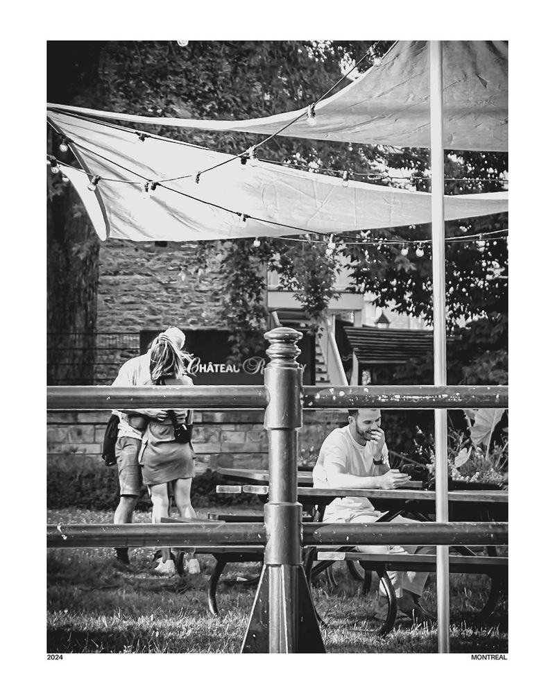 Black and white photo of a couple hugging in a park in Montreal, partially obscured by string lights and fabric overhead. A man sits alone at a picnic table in the foreground, appearing to observe or be deep in thought. The mood is cinematic and nostalgic.

