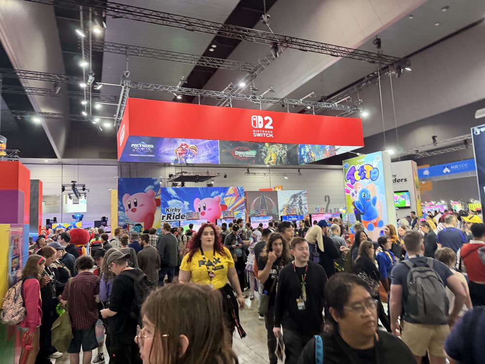 A large indoor gaming convention is crowded with attendees walking around and interacting with various booths. The central booth features a red banner with lighting rigs above and multiple game displays below. A giant poster of Kirby is visible, along with a vertical sign featuring a blue character labeled "TETTO." Attendees wear casual clothing and carry backpacks, and some are holding phones or event guides. Multiple screens and gaming setups are visible in the background, and the ceiling is lined with lights and structural beams.

Text: "Nintendo Switch", "Kirby Air Ride", "TETTO", "Metroid Dread"