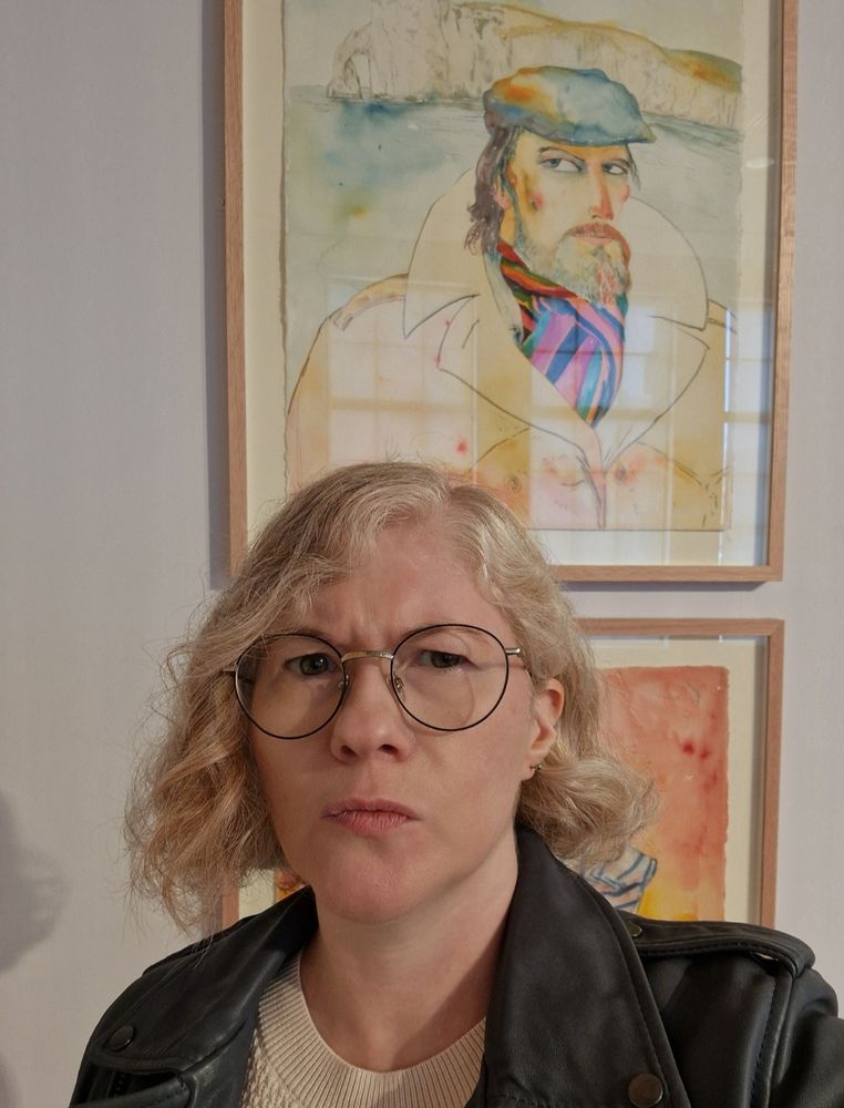 A white person with blonde curly hair and round glasses, wearing a dark leather jacket, standing in front of a pastel-coloured John Bellany drawing of a suspicious-eyed bearded man in a large trenchcoat