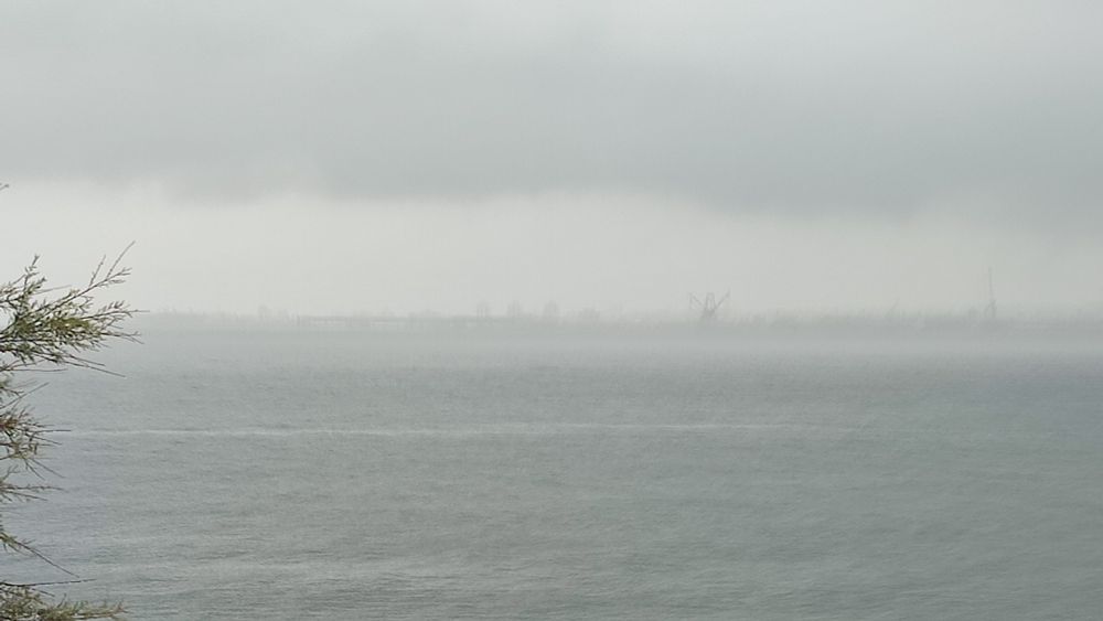 A bleak view of a grey sea under heavy clouds. Far on the horizon dark industrial shapes are barely visible through the rain.