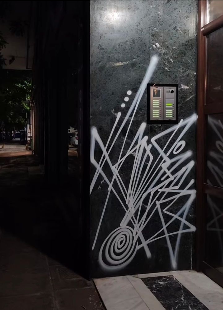 White graffiti on a black wall at night, with lots of pointy, jagged shapes and a spiral at the bottom. (photographer unknown)
