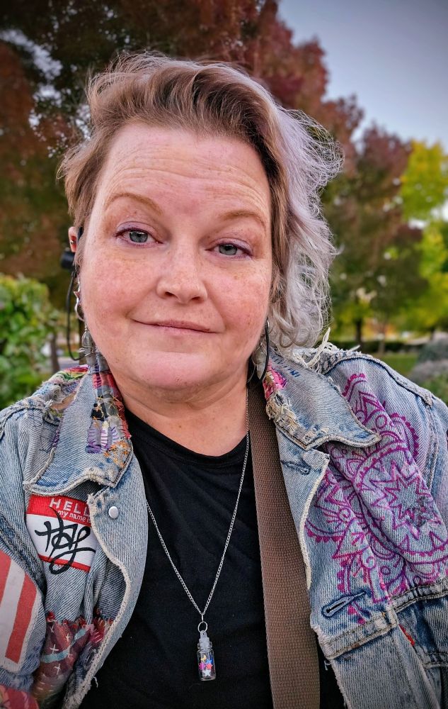 Selfie of an older white woman with short grey hair and a heavily patched & decorated jean jacket, in front of a group of trees.