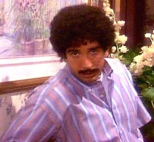um jovem Ross, personagem do seriado Friends, sustentando um bigode e um cabelo volumoso e cacheado, olhando para a câmera com olhar blasé, vestindo uma camiseta listrada em branco e rosé