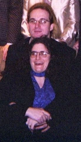 Ruby Tuesday sitting on a couch surrounded by family that has mostly been cropped out. Her husband sitting in front of her.