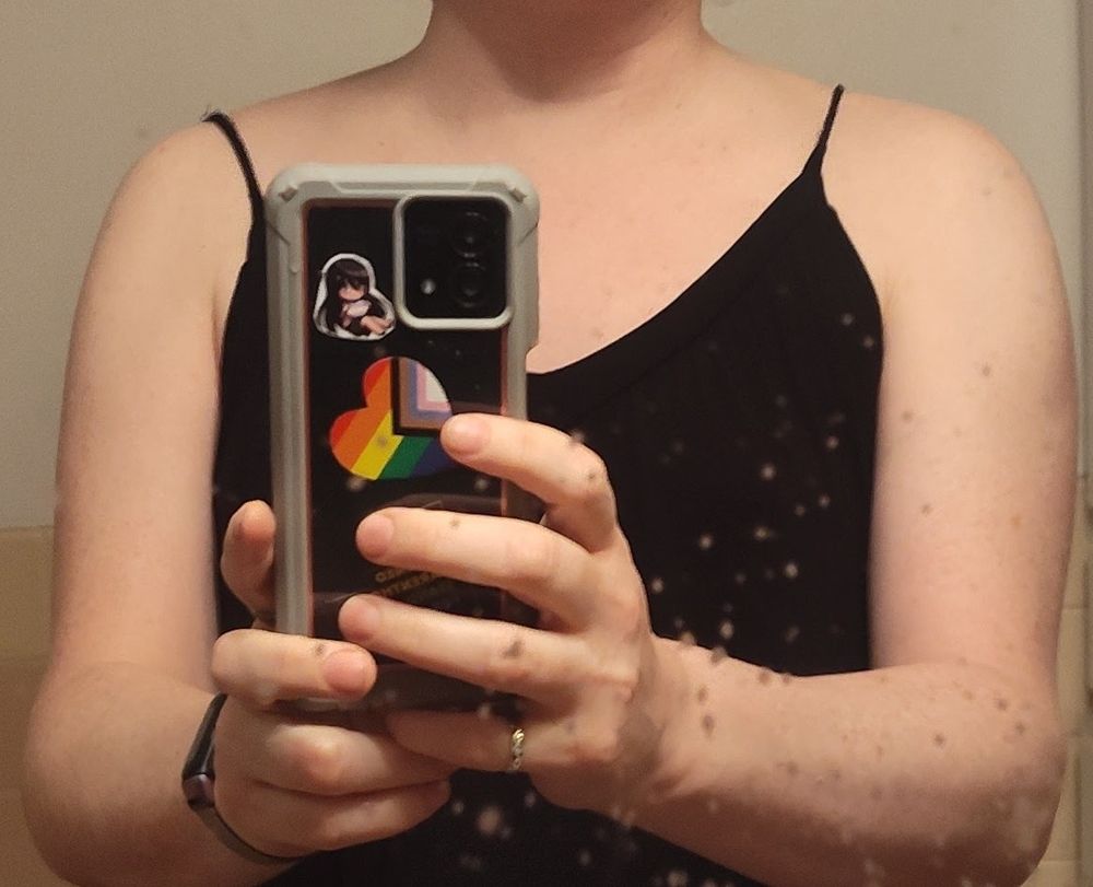 Ruby, from shoulders to elbows. She is wearing her black nightgown and holding her phone in front of her. The mirror needs to be cleaned.