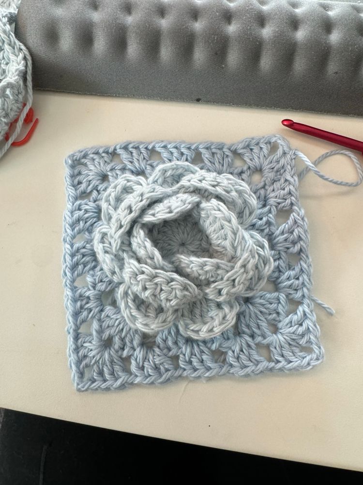 A light blue crochet granny square with a three dimensional flower in the center.