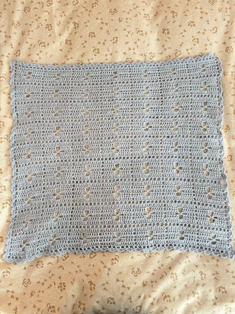 A square light blue crochet baby blanket laid flat onto a beige bedspread.