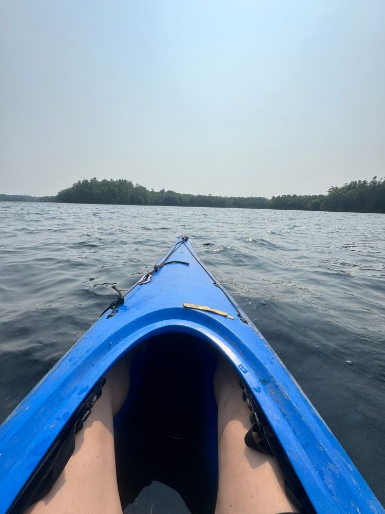 My legs tucked inside a blue kayak on Duckpuddle pond with the trees on the shore in the distance