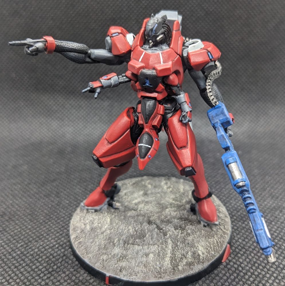 A miniature of a large combat robot, painted with red, black, and grey armour panels. Small blue lights dot regions on its chest and shoulders. Thin white lines run up its chest yowards a black head. It carries a large machine gun with a blue casing on its left arm. its right arm is pointing off screen in a menacing pose