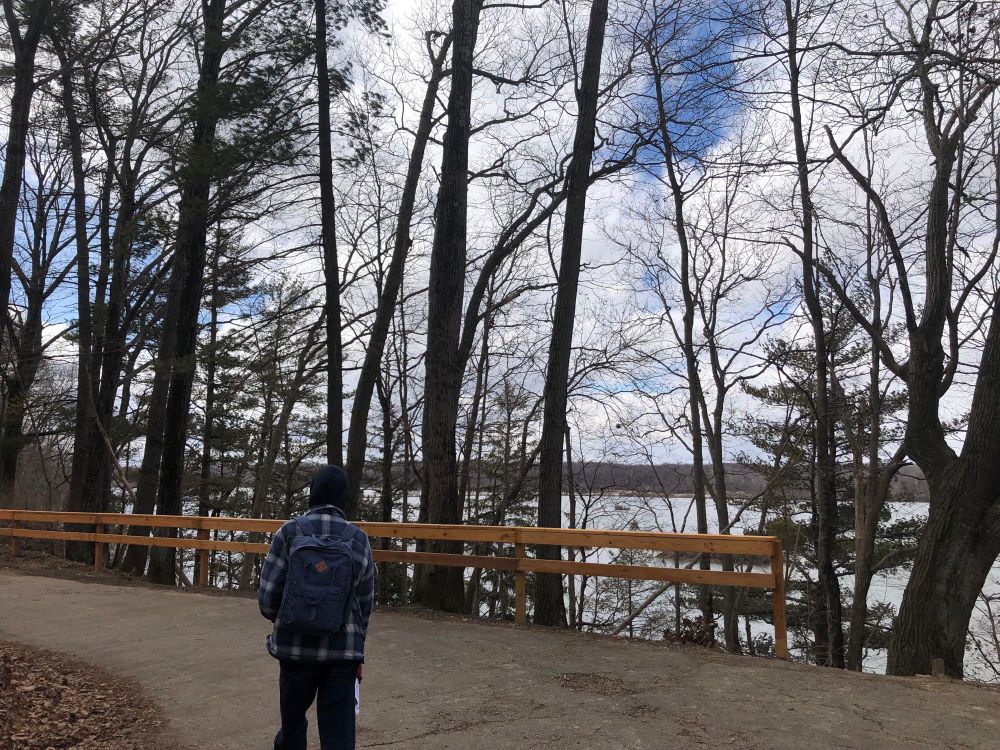 Cold kid with backpack hiking back to the car, passing a lake view under blue sky with fluffy white clouds.