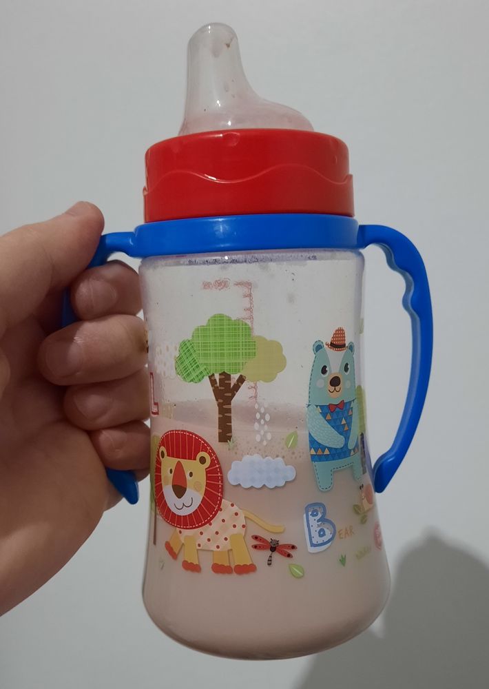 An image of a sippy cup filled halfway with chocolate milk. It has a drawing of a lion, a bear, and a tree on the plastic. It has a blue handle and a red top.