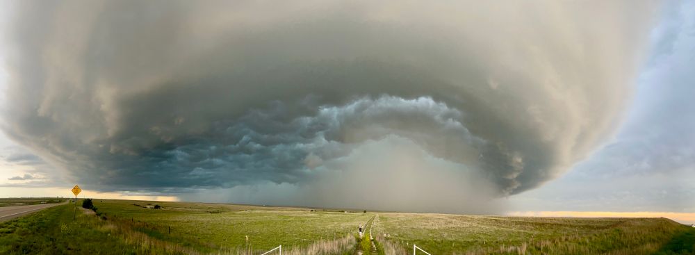 A panoramic view of a supercell, with a mothership-like structure. An undulating underside has a blue-green tinge, with a core of opaque heavy precipitation atop open grasslands 