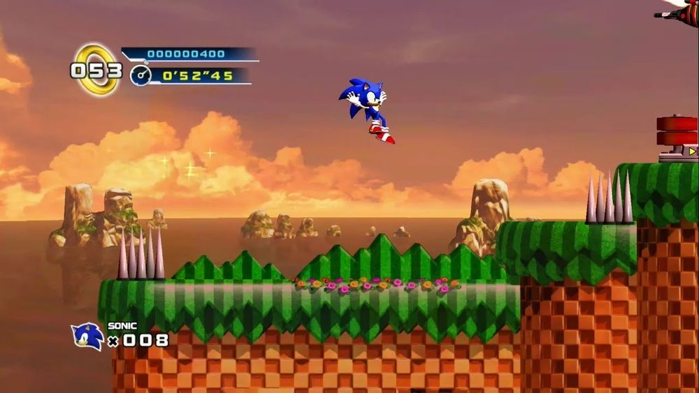Sonic in a mid-air jump in Splash Hill Zone Act 3 just above a grassy platform. With spikes on the left edge, more a little to the right, and a speed panel just above it to the right. Sunset background along with the ocean and rocks in the distance.