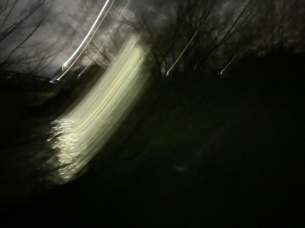 Night time picture of a river, with lights, the lights blurred and smeared across the image, which contains trees and water and bridges 