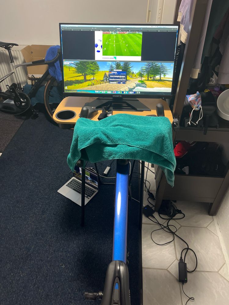 Zu sehen ist ein großer Monitor auf einem Beistelltisch vor dem Rennrad welches auf der Rolle aufgebaut ist.
Auf dem Monitor ist die Bundesligakonferenz sowie das Programm TrainingsVirtual zu sehen.