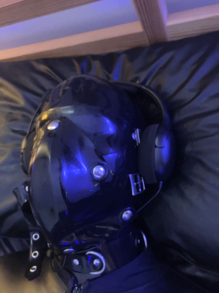 A gimp in black rubber. Close up of his head lying on a black rubber bed sheet. A black rubber catsuit codpiece upside down over his mouth and nose. 