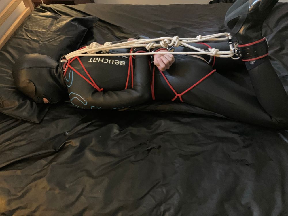 A young skinny white man wearing neoprene triathlon wetsuit, booties and hood. He is in rope bondage with a metal hook in his ass in a ‘hogtie’ like position on his front. 
