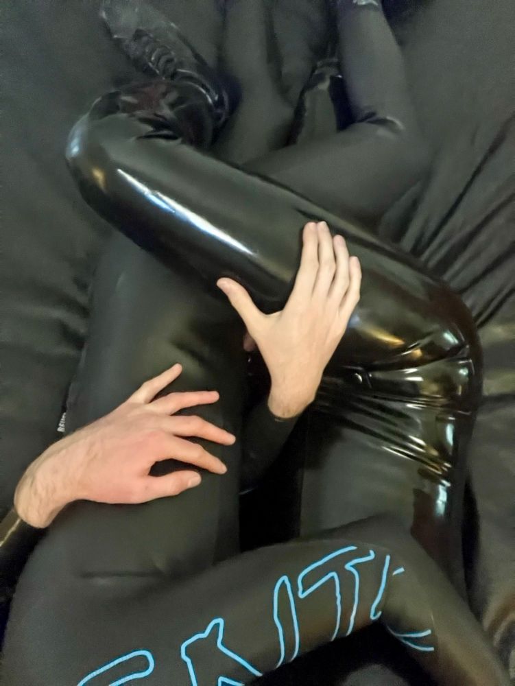 Neck-down selfie of two skinny white men cuddling on a bed with legs wrapped around each other. One is wearing a triathlon wetsuit, the other a black latex catsuit. 