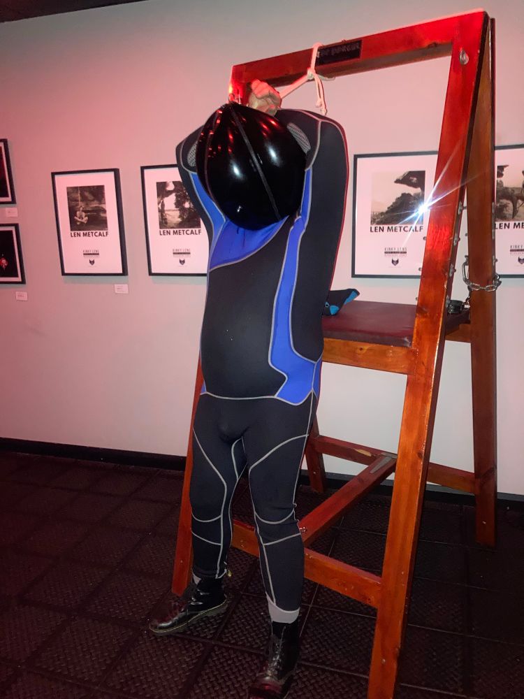 A tall man wearing a blue and black wetsuit and leather boots. His head is concealed by an inflatable rubber ball hood. He is stood in front of a wooden bondage frame with his hands tied above and behind his head. 