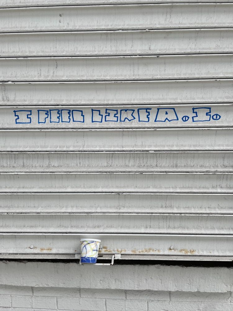 Grafitti says in block letters: "I feel like A.I." The dots look like eyes. Beneath it, an abandoned coffee cup.