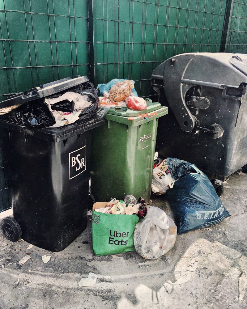 Szene mit mehreren Mülltonnen auf und vor denen etliche Müllbeutel abgelegt sind, einer davon mit der Aufschrift „Uber Eats“.