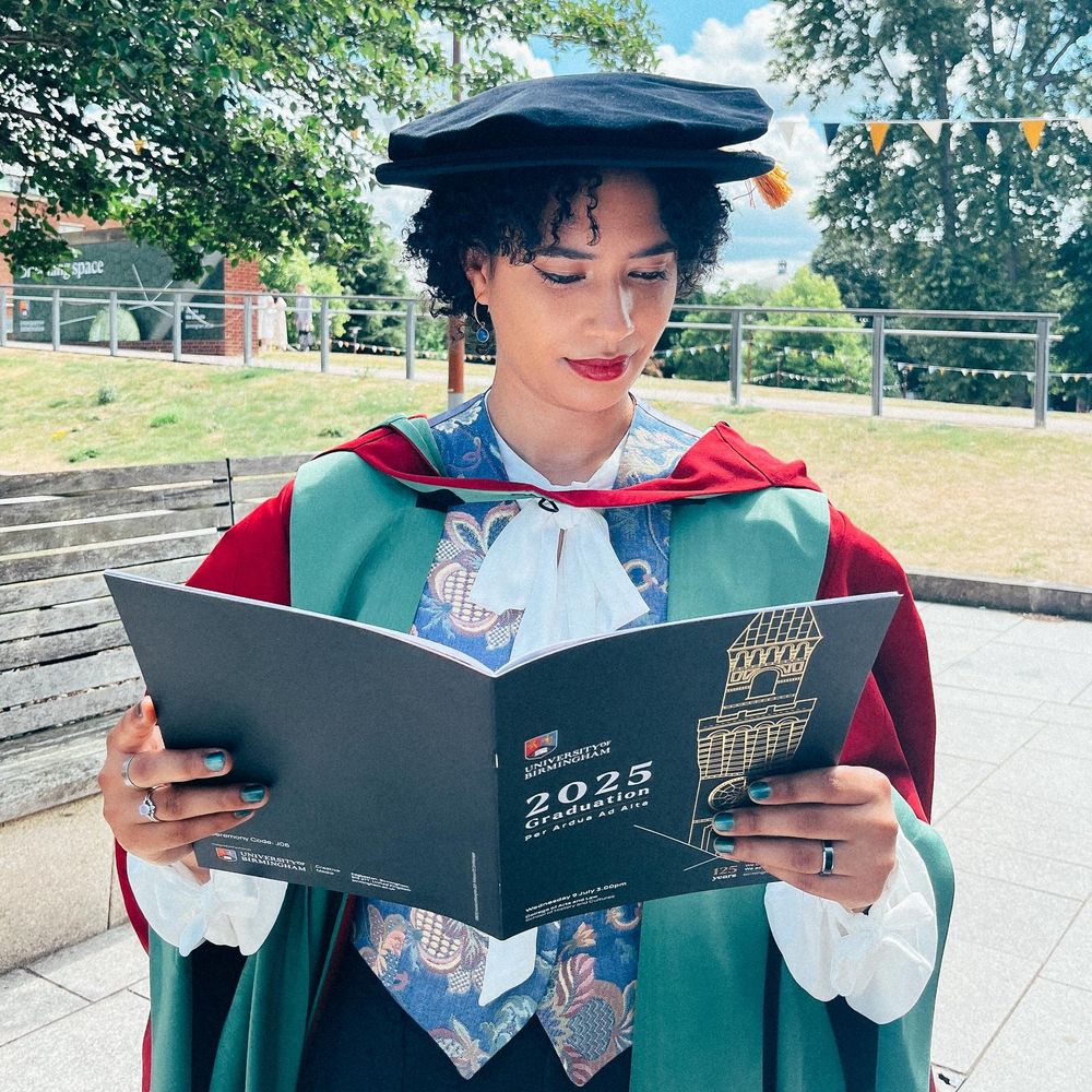 Annabelle is wearing doctoral graduation robes and reading the University of Birmingham graduation programme.