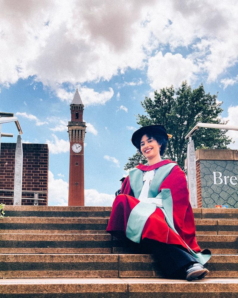 Annabelle is sat on some steps in her doctoral graduation robes with the University of Birmingham clocktower, 'Old Joe', in the background.