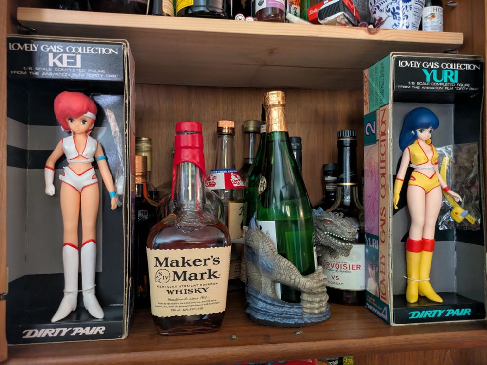 kei and yuri figures from the lovely gals collection flanking our liquor cabinet. the taco bell 98 godzilla cup holder can be seen clutching a bottle of nice sake between them