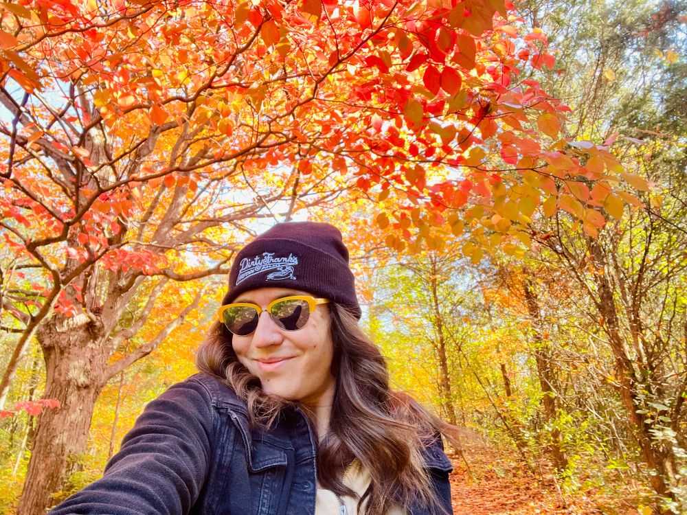 a smiling brunette wearing a black hat and black jacket in front of a multitude of colorful leaves