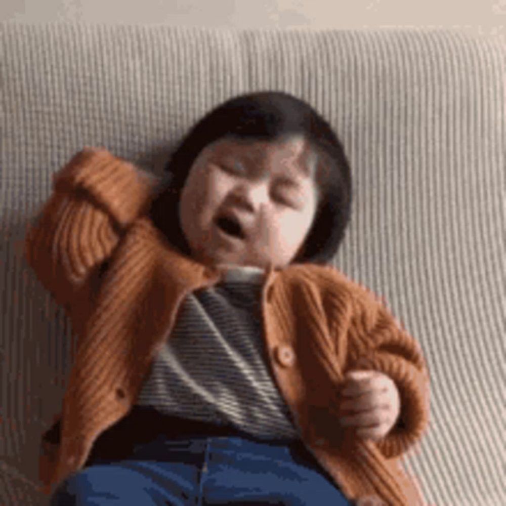 a baby is yawning while sitting on a couch wearing a sweater .